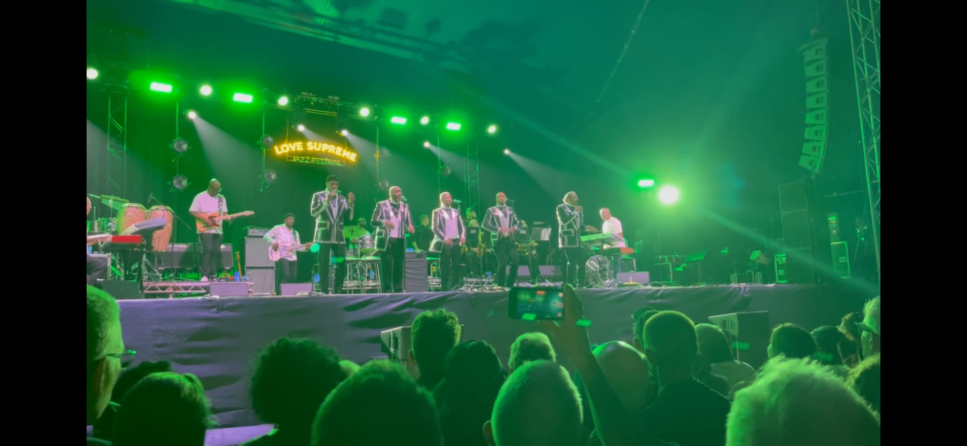 The Temptations performing on the South Downs tent stage at the Love Supreme Jazz Festival, bathed in green light while a packed crowd sings along.