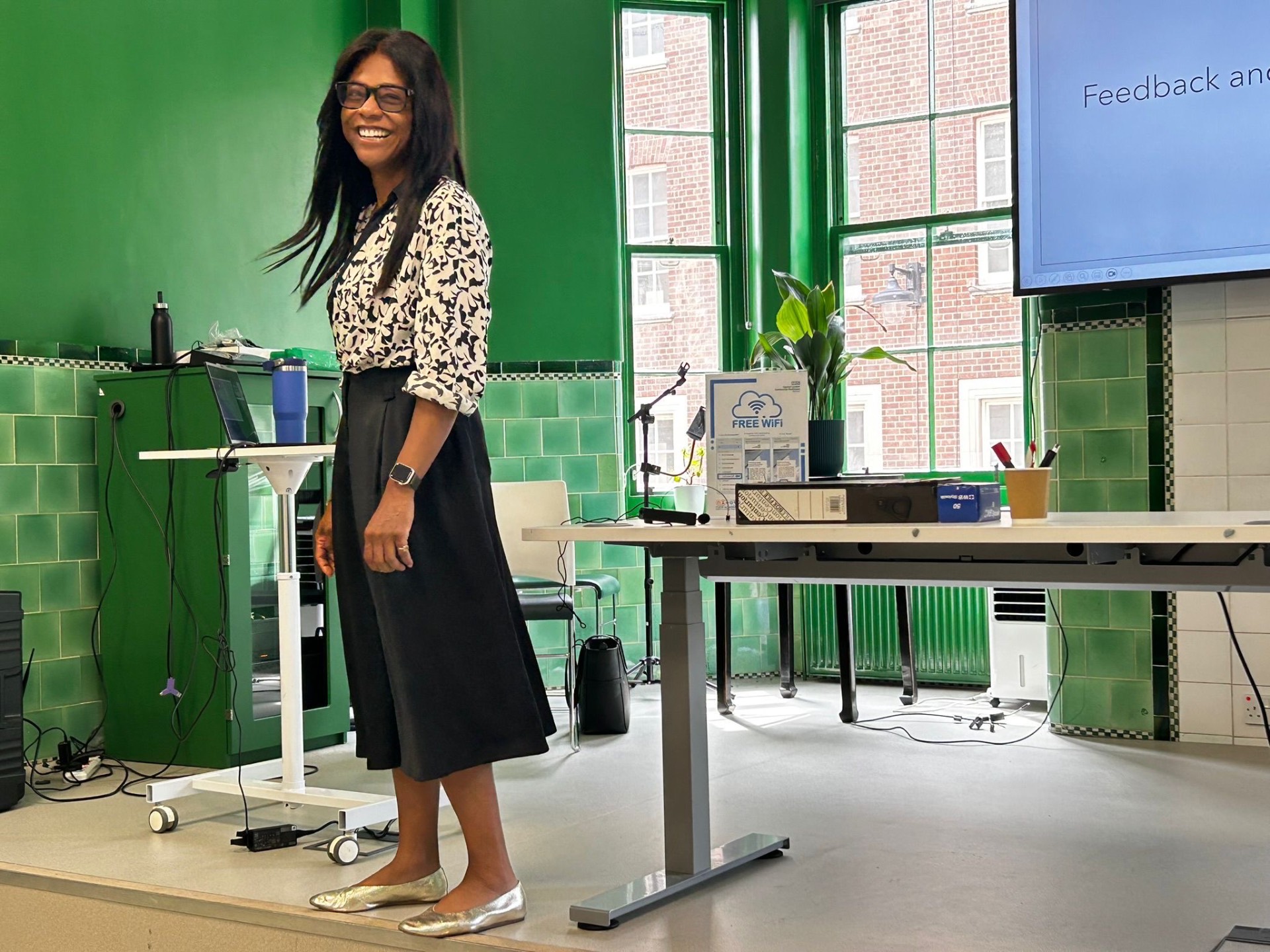 Janine La Rosa smiling while leading a session on joy and feedback in a bright green room, standing near a screen and a desk with a laptop and microphone.