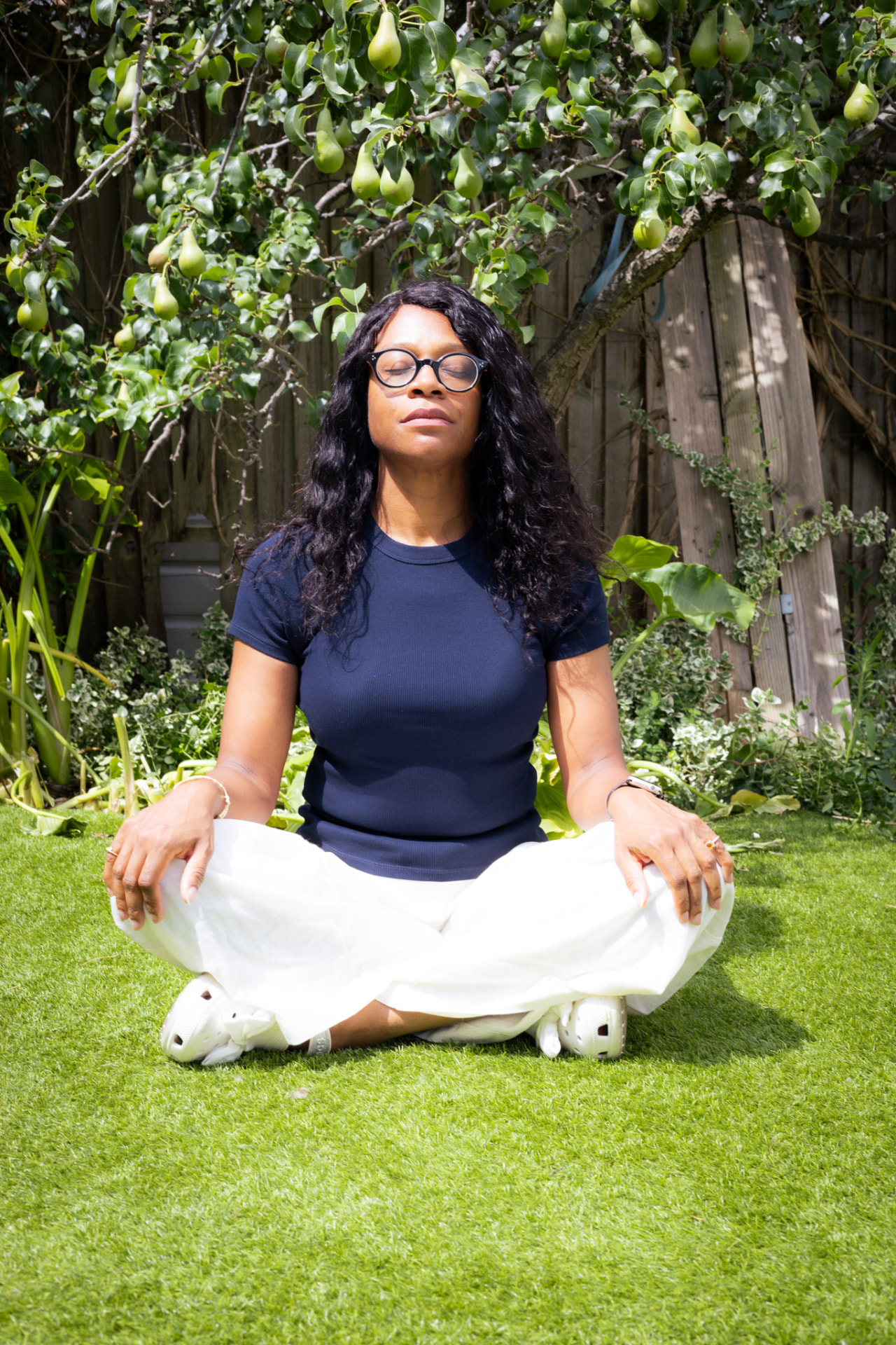 Janine La Rosa sitting cross-legged on the grass beneath a pear tree laden with fruit, eyes closed, hands resting open on her knees in quiet practice.