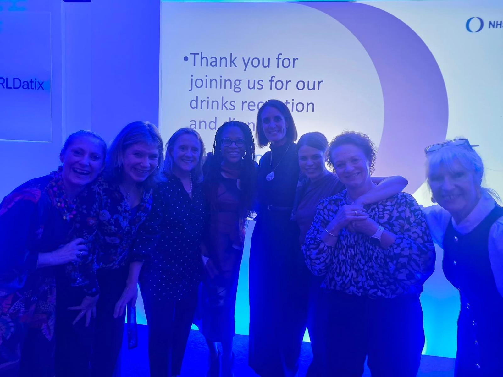 Janine La Rosa and fellow Chief People Officer colleagues standing together and smiling at an evening reception, under blue lighting in front of a welcome screen.