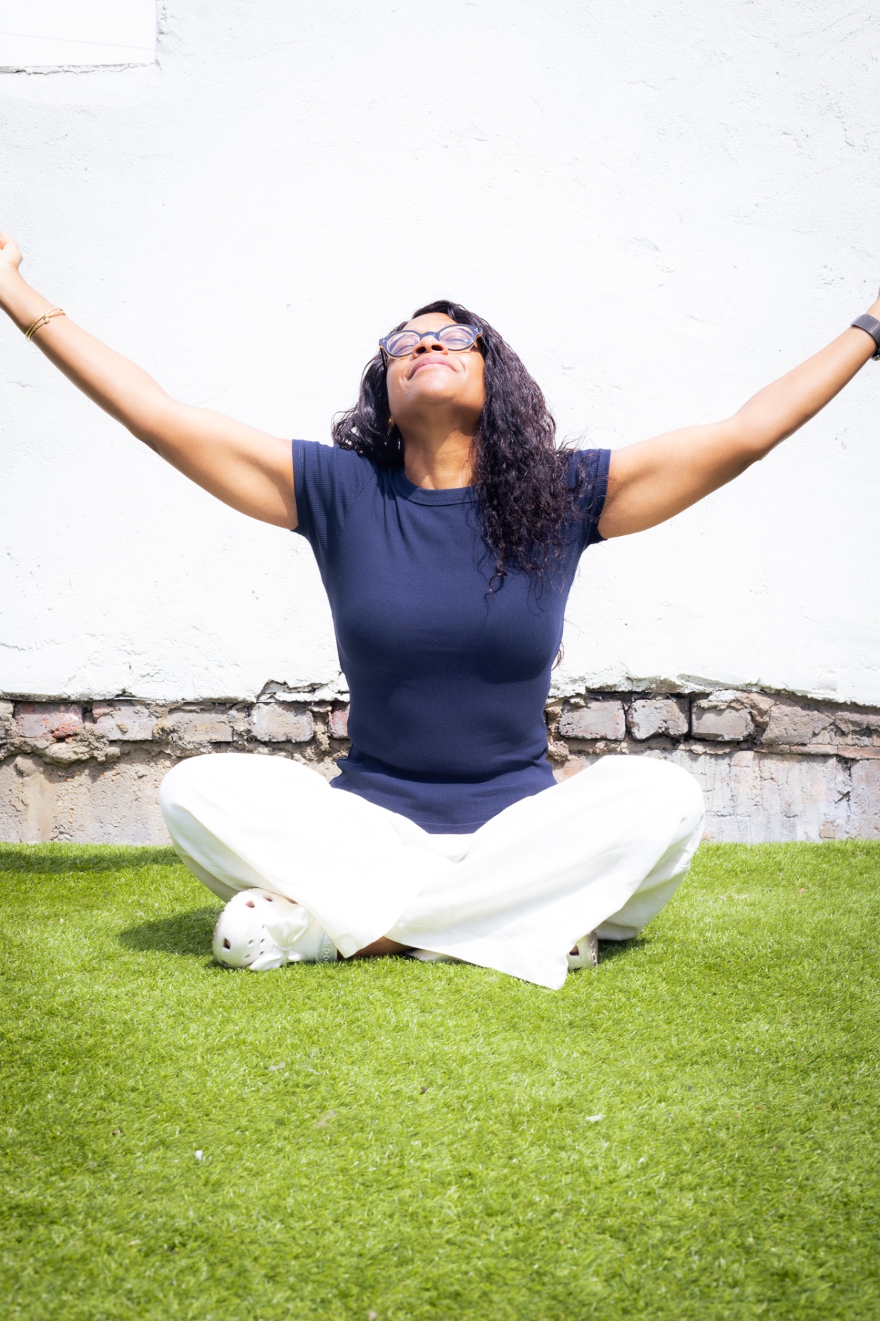 Janine La Rosa sitting cross-legged on the grass with arms open and face tilted toward the sky, eyes closed in a moment of mindful, embodied pause.
